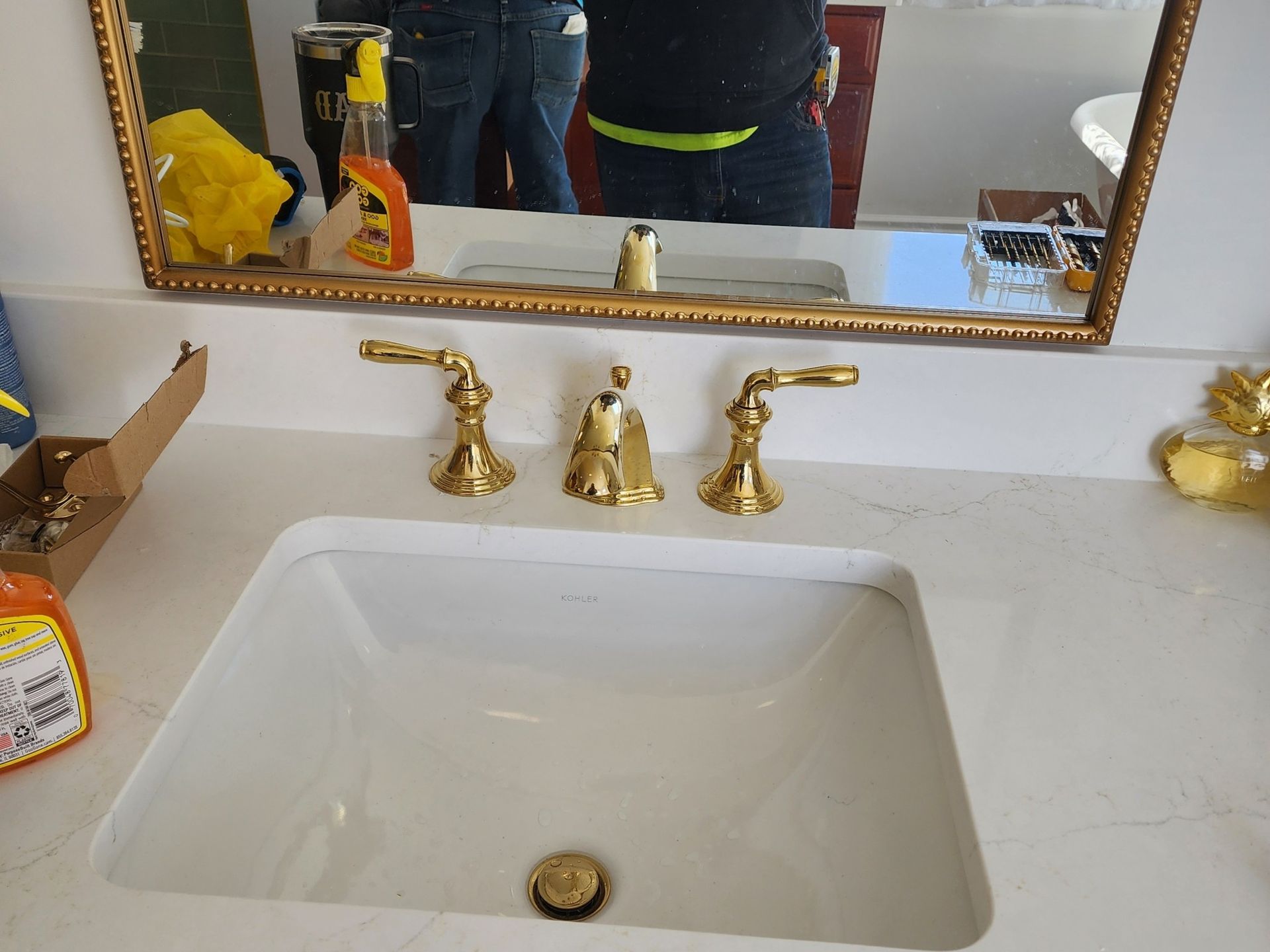 Gold faucet and white sink in a bathroom, with a mirror reflecting a person.