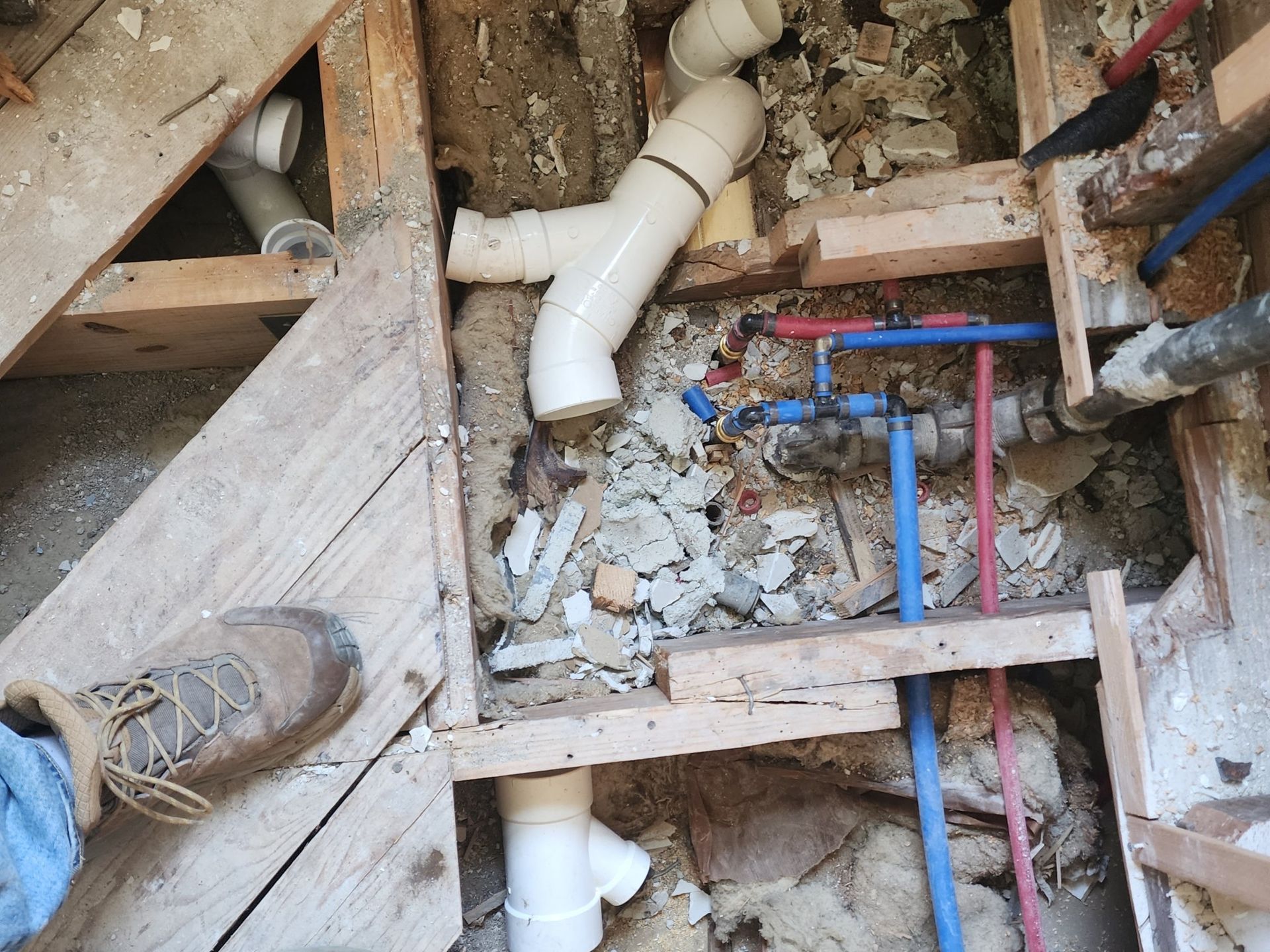 Plumbing pipes and fittings exposed in a floor opening, including drain, water lines, and a worker's boot.