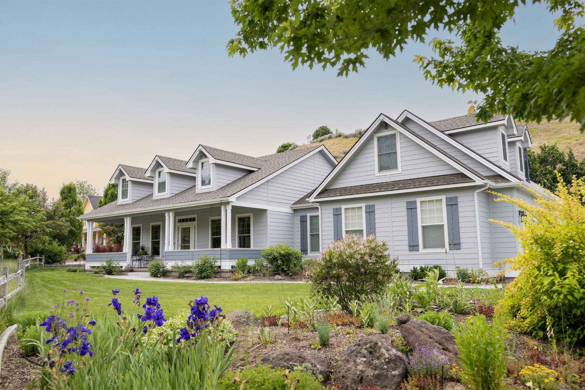 Light blue house with a covered porch, dormers, and a landscaped front yard with flowers.