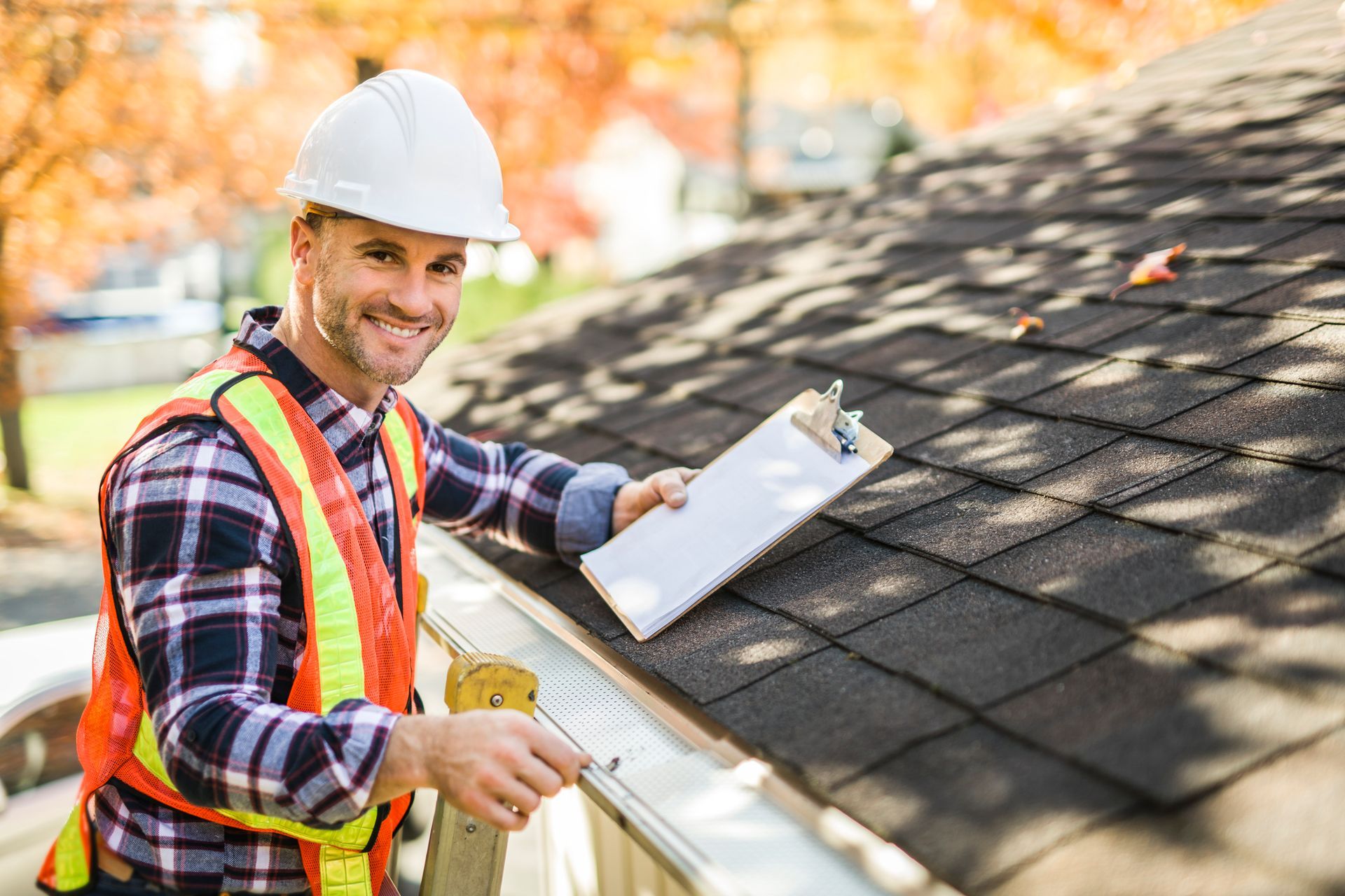 Man in hard hat and safety vest inspecting a roof, holding a clipboard and smiling.