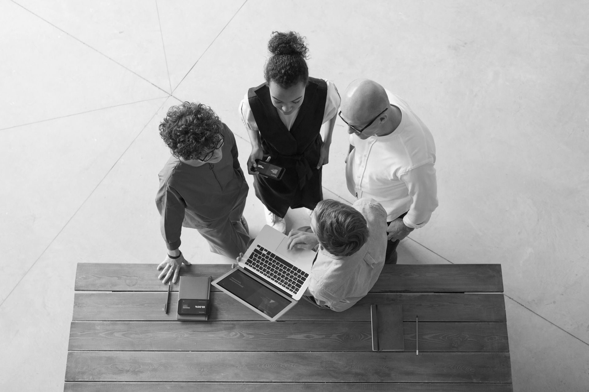 Four people standing around a wooden table, looking at a laptop and small device in a modern, light-filled room.