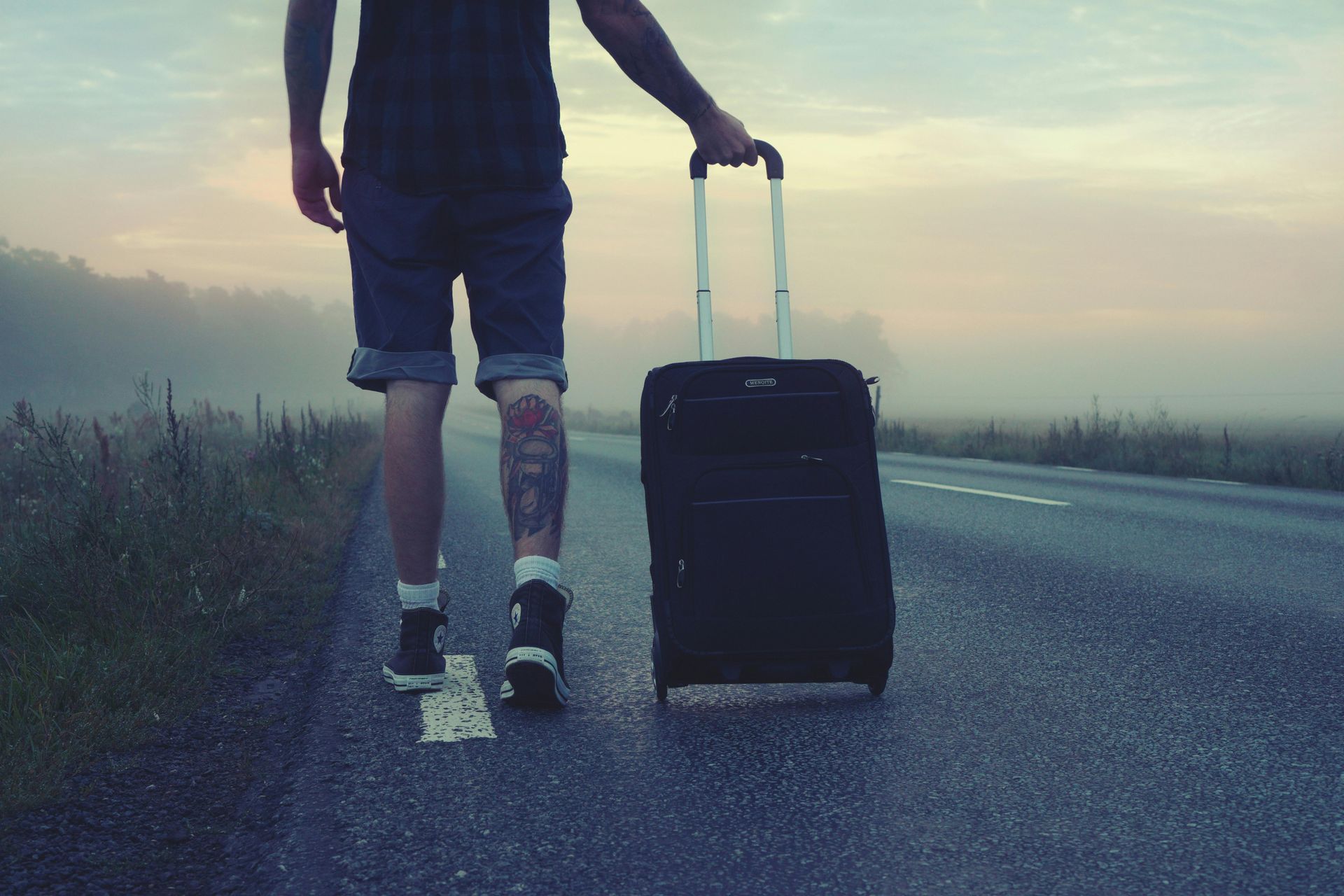 Man with tattoos rolling a suitcase down a road, early morning with fog.
