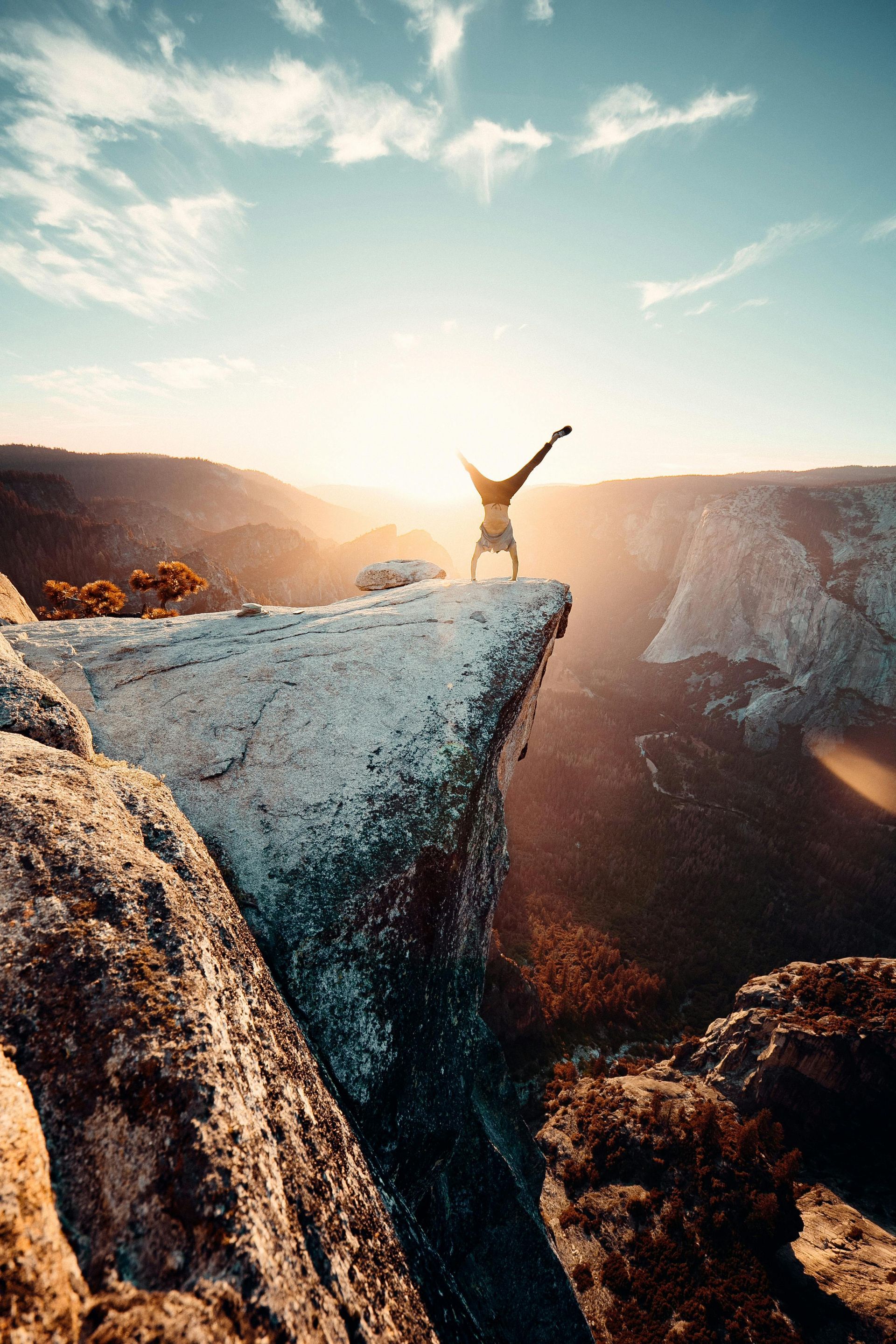 Person doing a handstand on a cliff edge, silhouetted against the sunset over a mountain range.