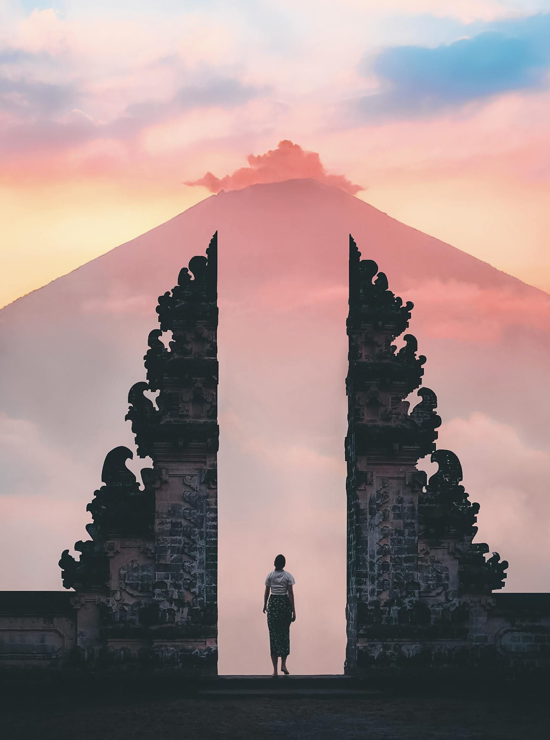 Woman standing in ornate gate, facing mountain peak at sunset. Pink and orange hues.