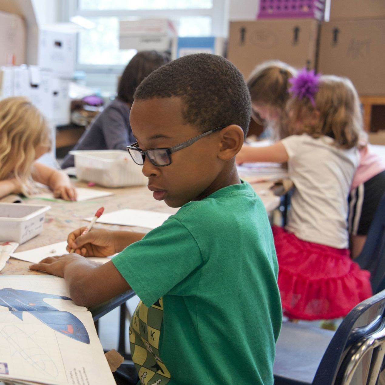 A young boy wearing glasses sits at a table with other children