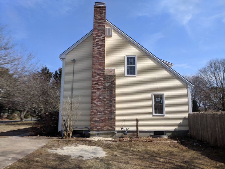 Side view of a house with light tan siding, a brick chimney, and two windows. Sunny day.