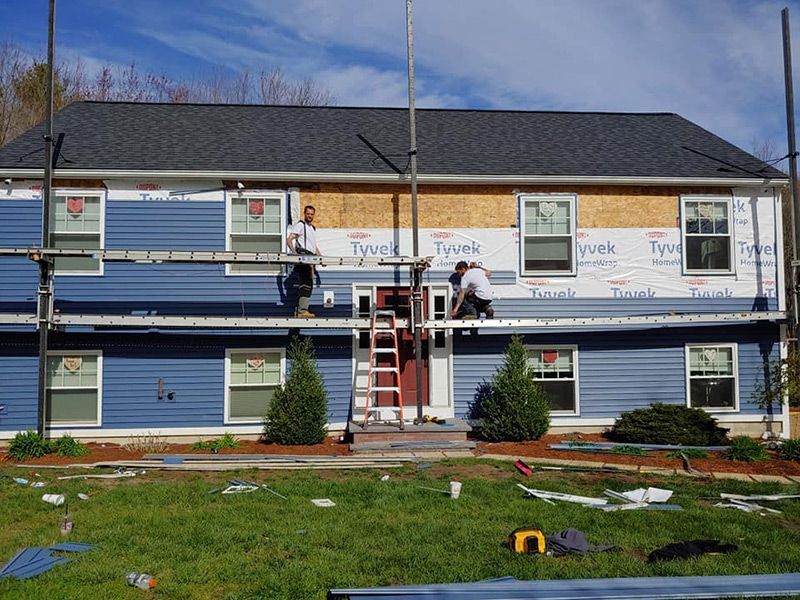 Two-story house under construction with blue siding. Workers on scaffolding install siding; the sky is blue and the lawn is green.