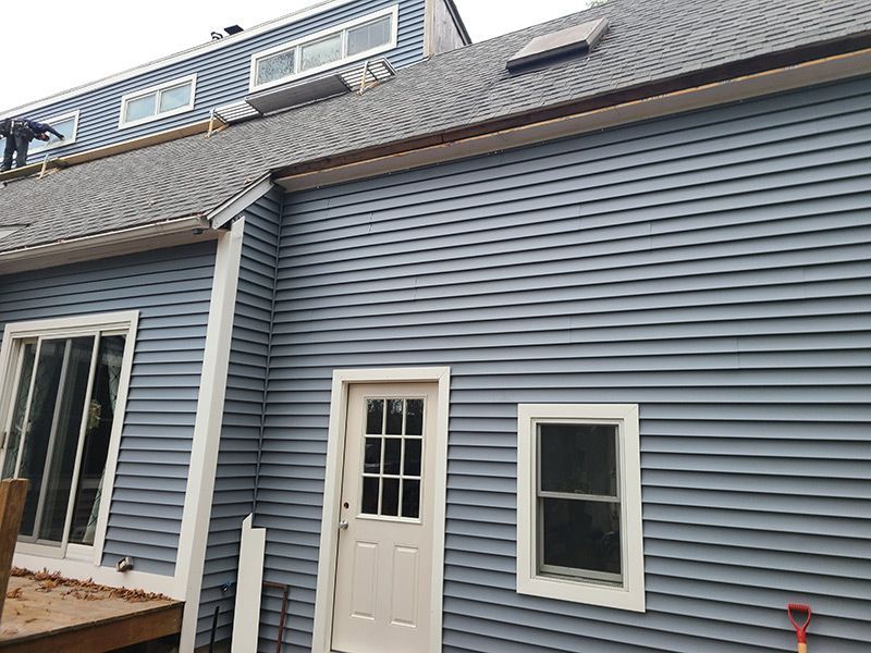 Blue siding on a house with a white door and windows. Gray roof and skylight visible.