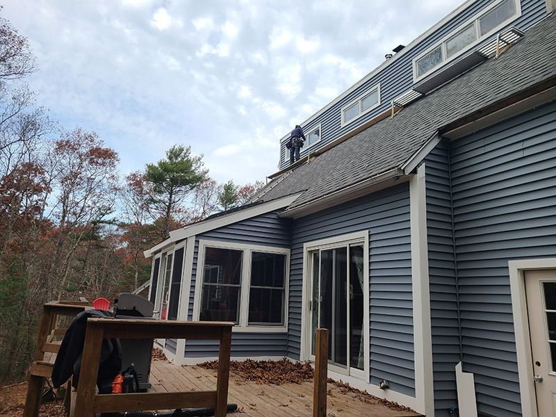 Person on rooftop of a blue-sided house, likely installing something. Deck in foreground, cloudy sky.