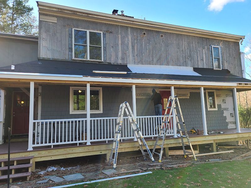 House under construction with worker on a ladder, installing siding on a porch; gray and white paint.