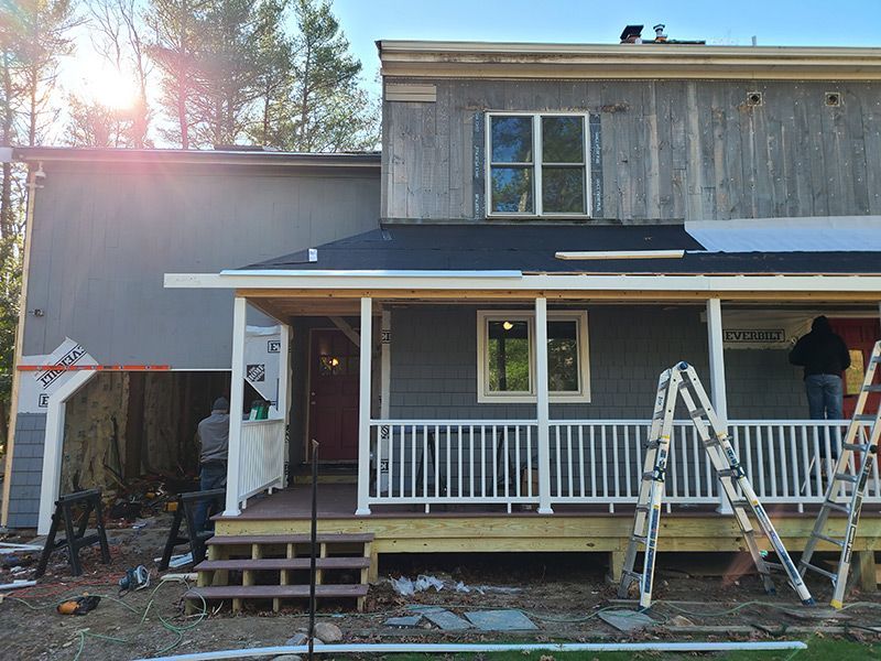 Home exterior under construction. Grey siding, porch with white railing, workers present.