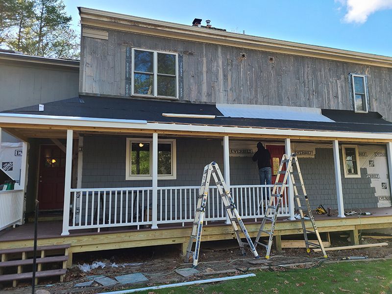 Exterior of a house under renovation, with siding and roof work in progress. A person is working on the porch.
