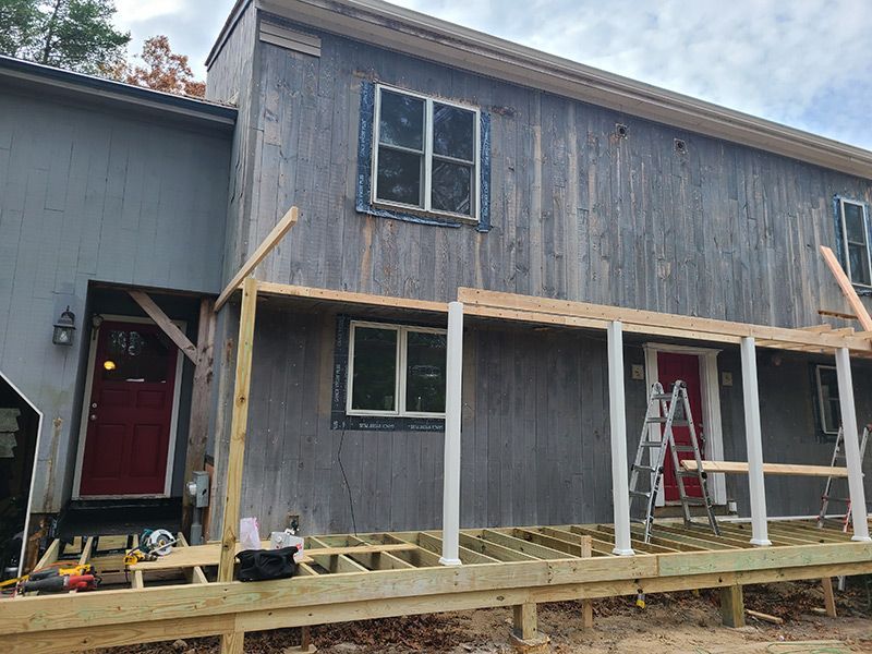 Exterior view of a house undergoing porch construction. Wooden frame, white columns, red doors, gray siding.