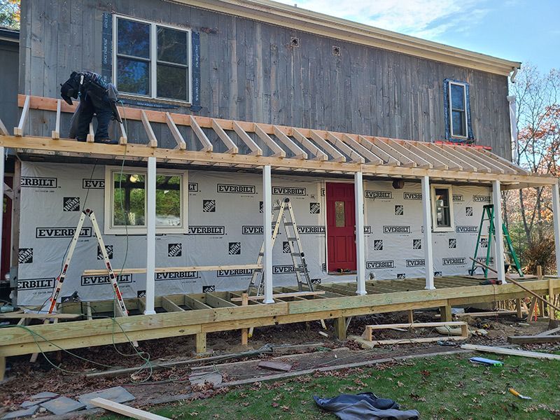 Construction of a porch with a worker on the roof. New support beams, white columns, and red door.