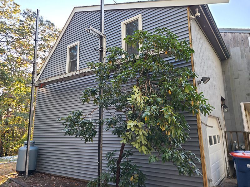 Gray-sided building with two windows and a garage door, with a street sign and a leafy tree in front.