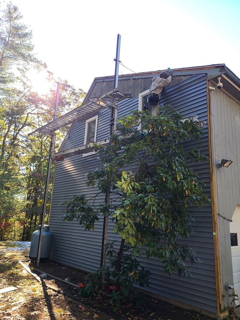 Side view of a blue-gray house with a chimney and ladder. A tree is in front. Sunny outdoor setting.
