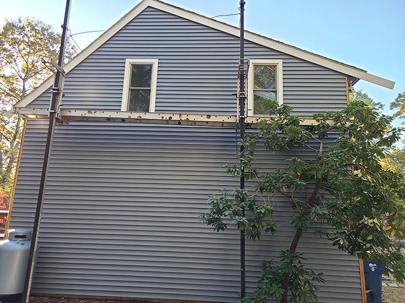 Grey house with corrugated siding, two windows, scaffolding, and a tree.
