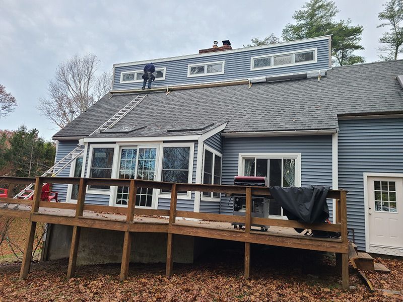 Person on a roof working on dormers, with a ladder and wooden deck in front.
