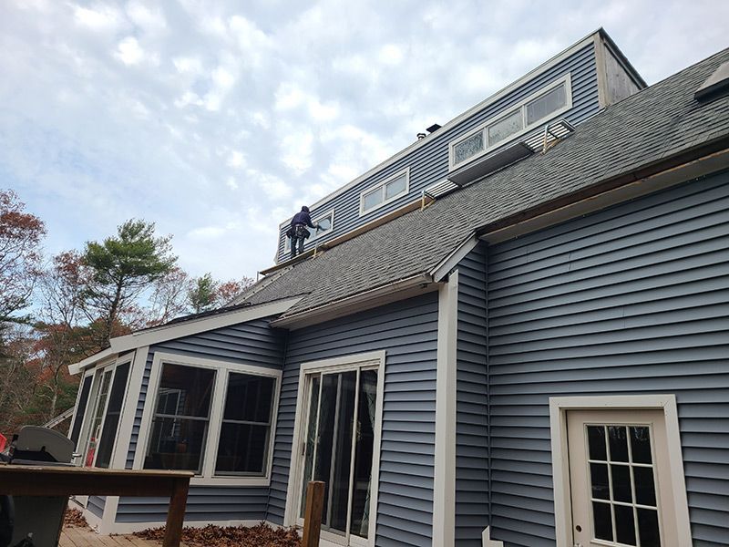 Person on a roof working on a blue-sided house with a cloudy sky overhead.