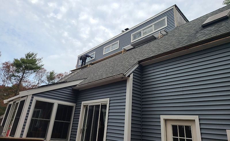 Person working on a dark gray shingled roof of a blue house with white trim against a cloudy sky.