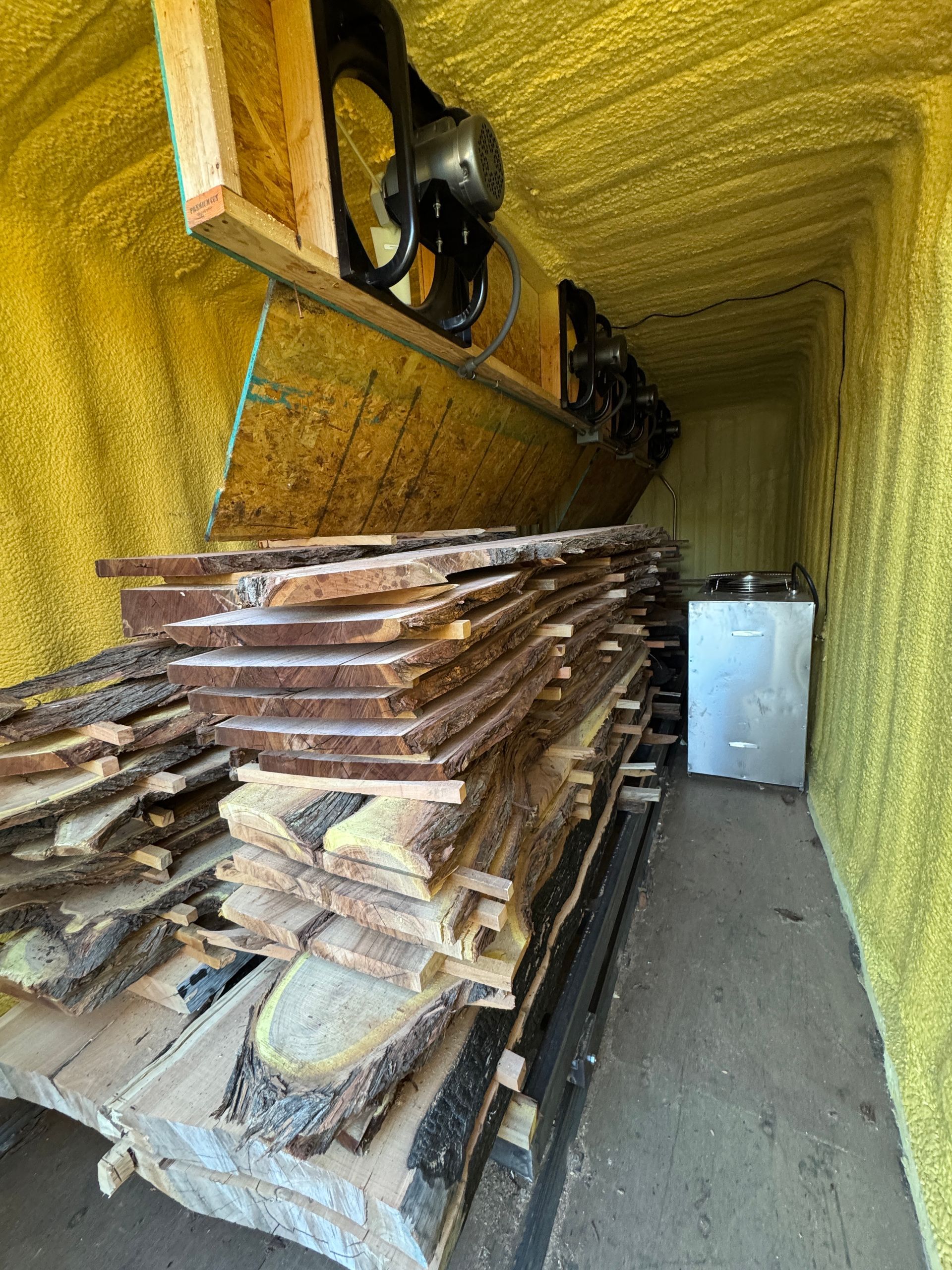 Wood planks stacked inside a yellow-insulated enclosure, fan overhead, light-colored appliance in the distance.