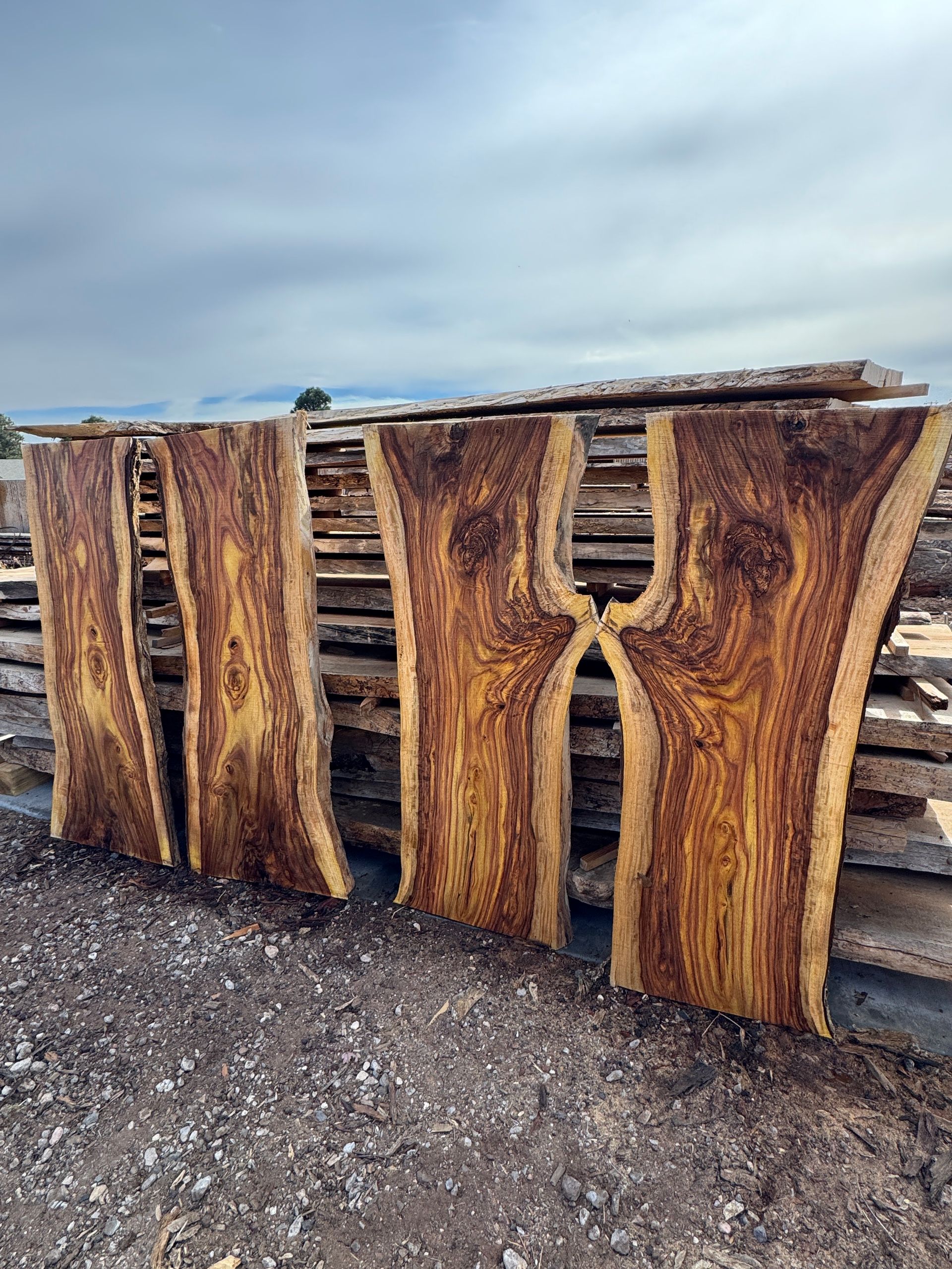 Live edge wood slabs, brown and yellow, stacked outdoors against a cloudy sky.