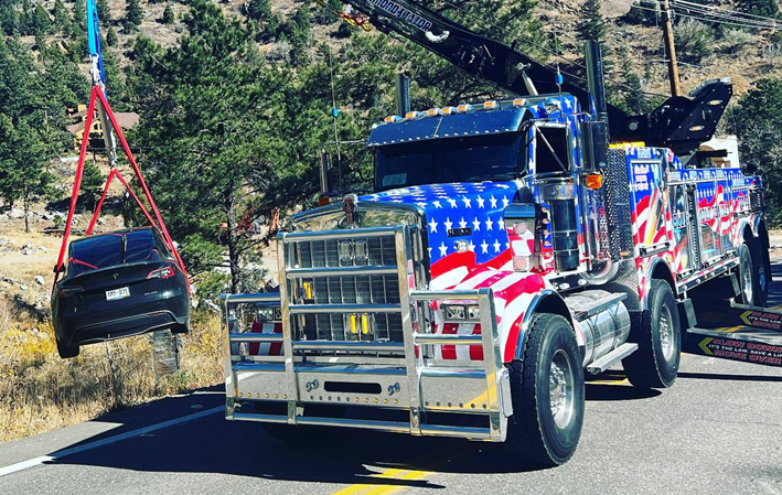 Tow truck hoisting a black car, decorated with American flag, on a mountain road.