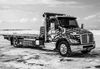 Black and white tow truck with American flag design parked on snow-covered ground under a cloudy sky.