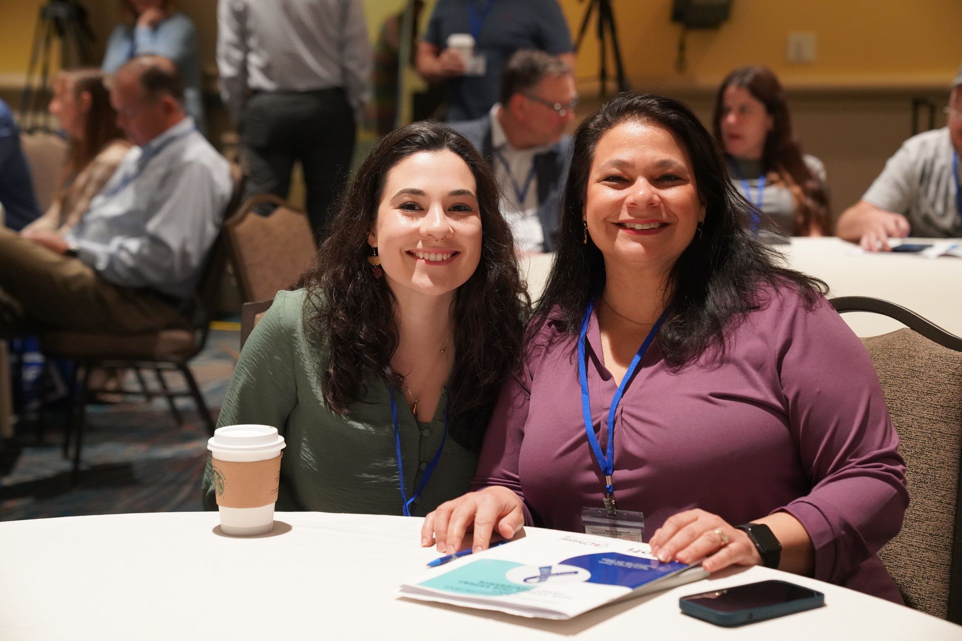 Two women smiling, seated at a table with coffee and a pamphlet in a conference setting.