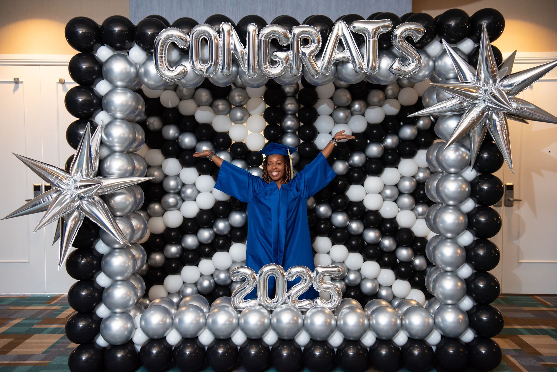 Person in blue graduation gown with arms raised, framed by balloons reading