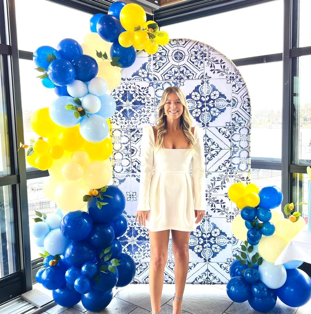 A woman in a white dress is standing in front of a blue and yellow balloon arch.