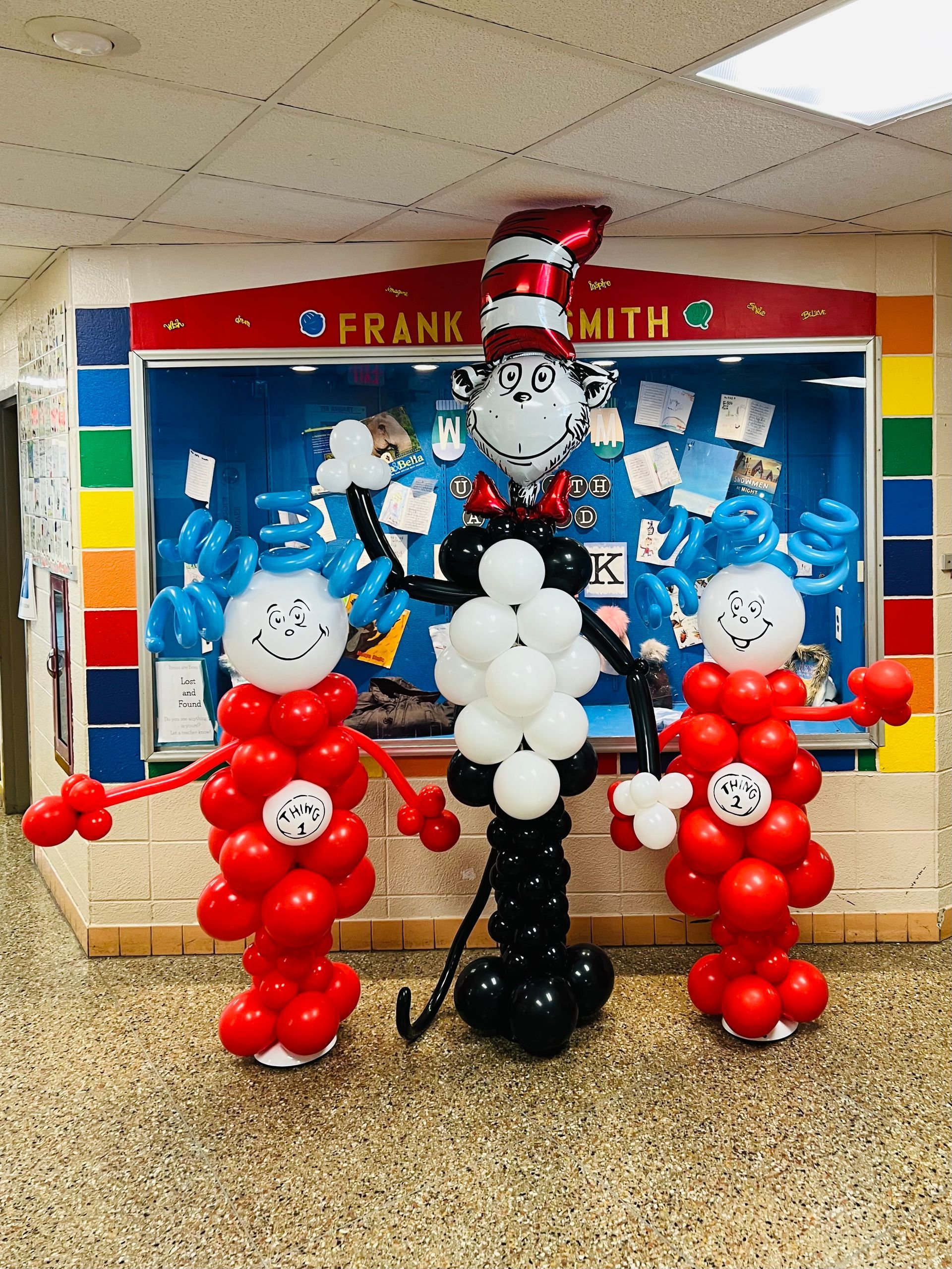 The cat in the hat is made out of balloons in front of a bulletin board.