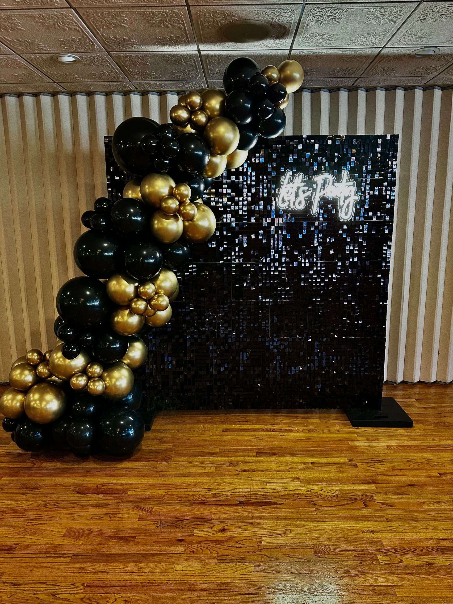 A black and gold balloon arch is sitting in front of a black sequined wall.