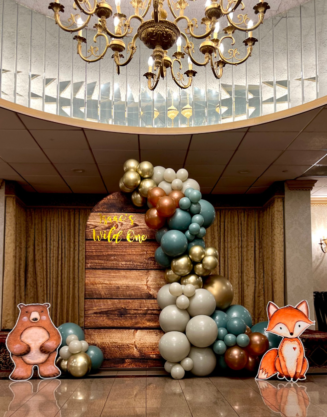 Birthday party decoration with balloon arch and woodland animal cutouts in front of a wood-textured backdrop.