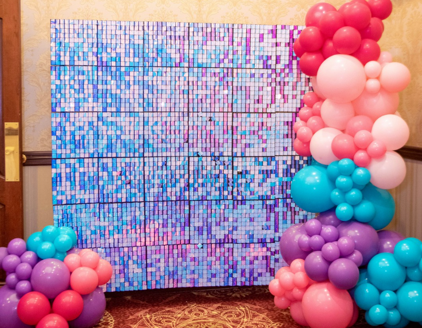 Colorful balloon arrangements next to a mosaic wall display. Balloons are pink, purple, and blue. Wall features various shades of blue and purple tiles.
