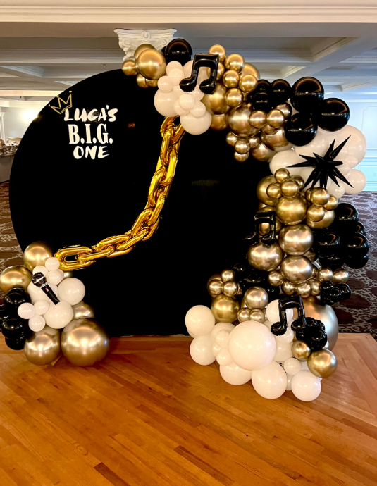 Black, gold, and white balloon arch with music notes and a gold chain on a black backdrop that says, “Luca’s B.I.G. One”.