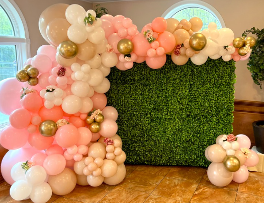 Balloon arch and floral arrangement in shades of pink, peach, gold, and white against a green hedge wall.