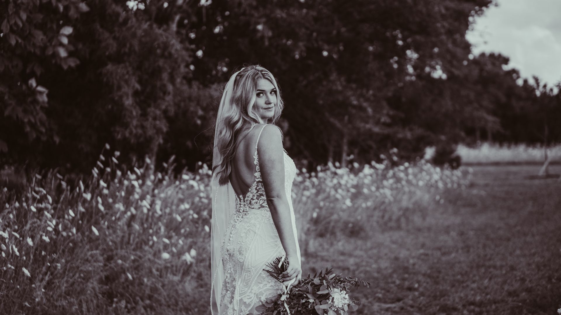 a bride in a wedding dress is standing in a field holding a bouquet of flowers .