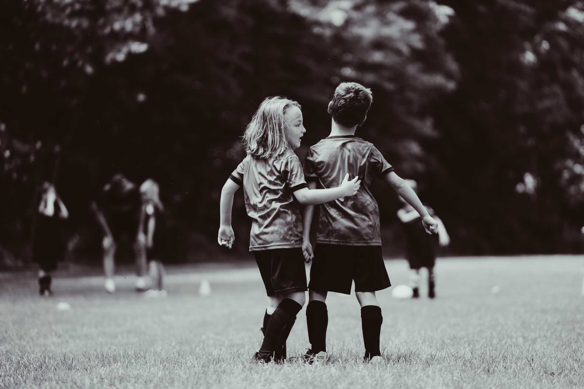 Two young soccer players with arms around each other, smiling, on a field; teammates.