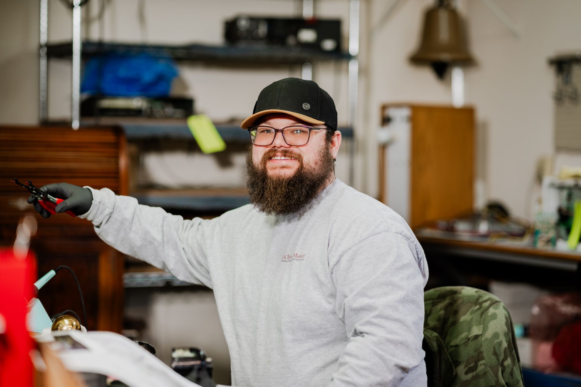 Man in a cap and beard holding pliers, smiling. Inside a workshop with shelving and objects.