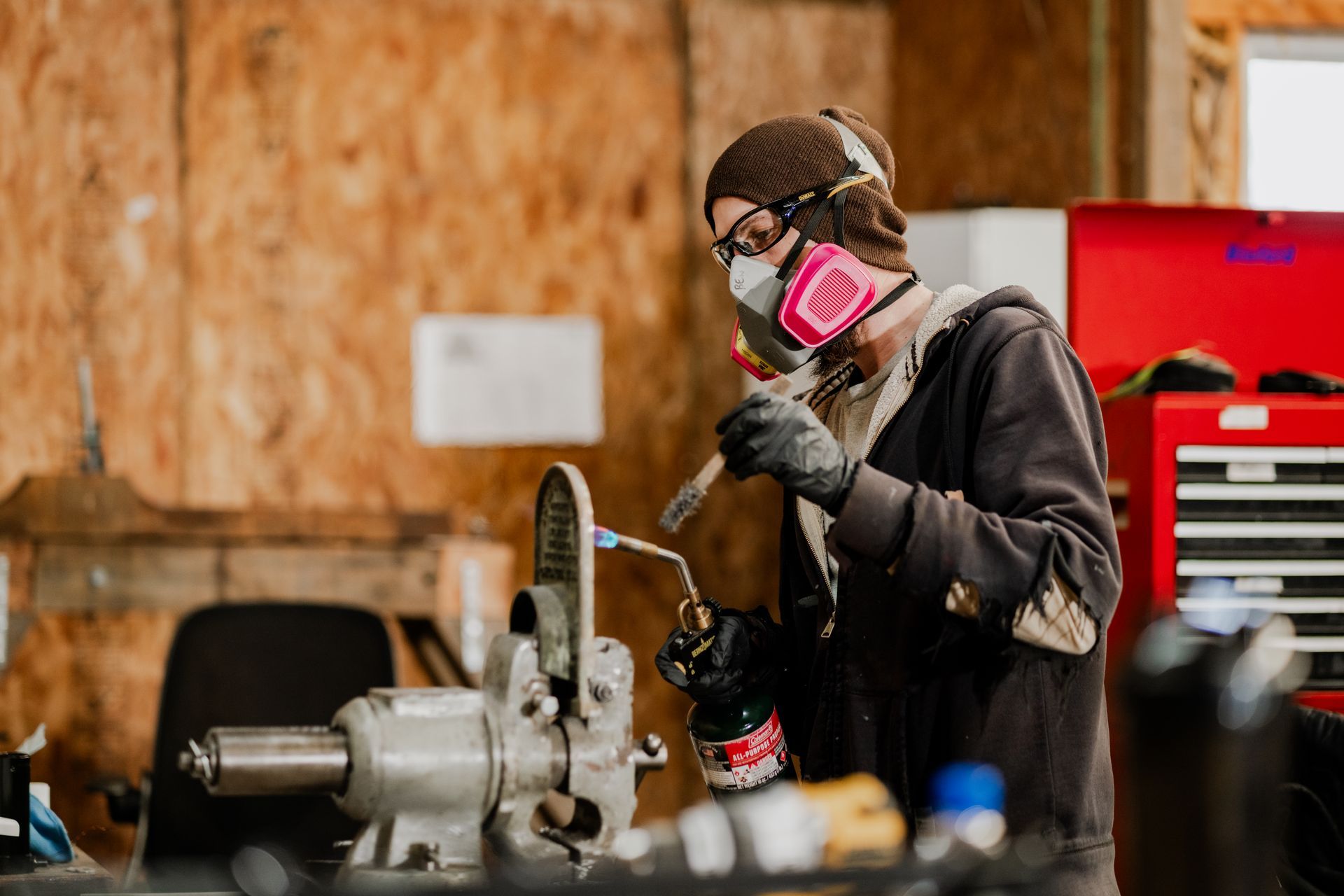 Person in workshop using a torch with safety mask, working on a metal object. Red toolbox in background.