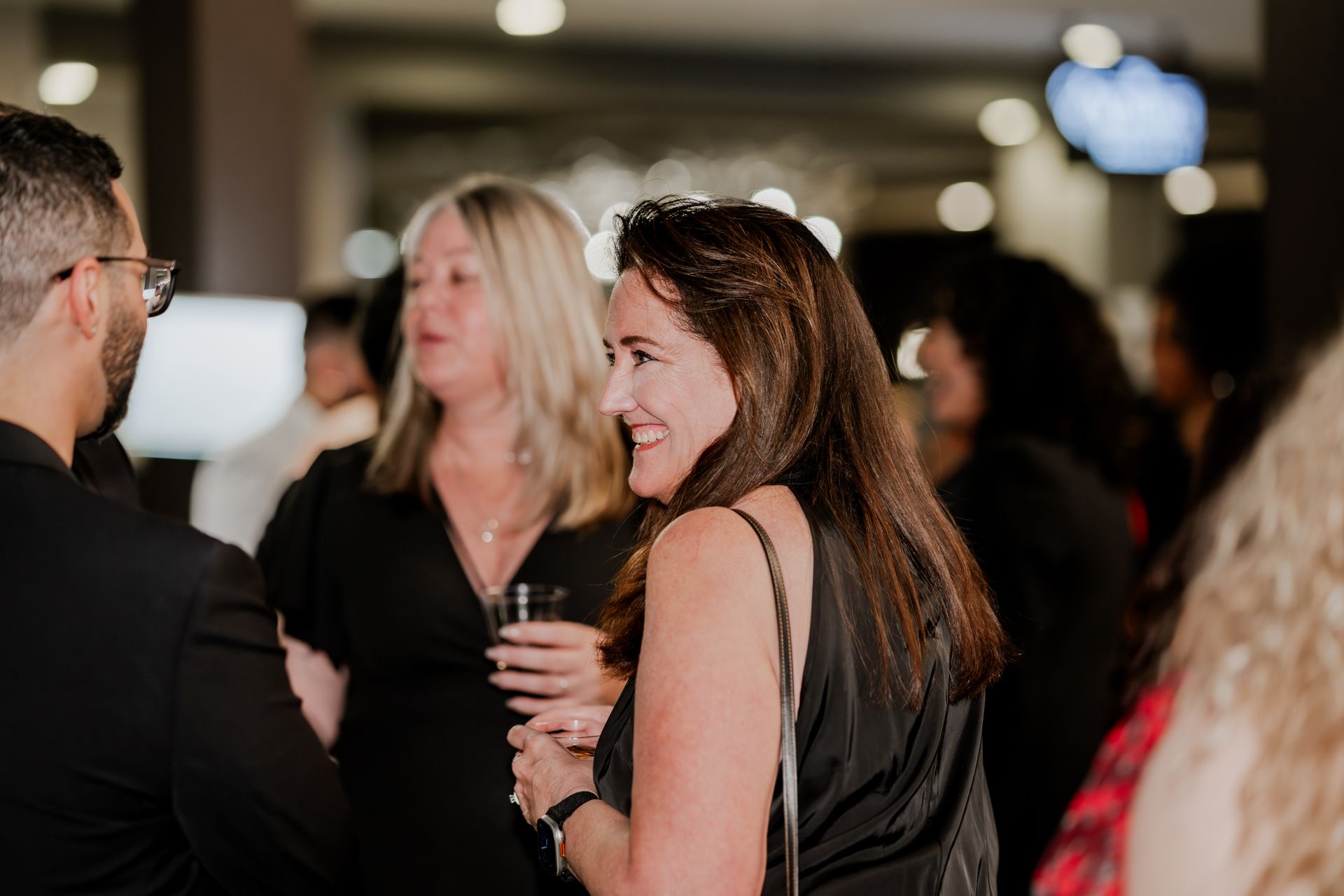 People in formal attire talk and smile at an indoor professional networking event with warm lighting.