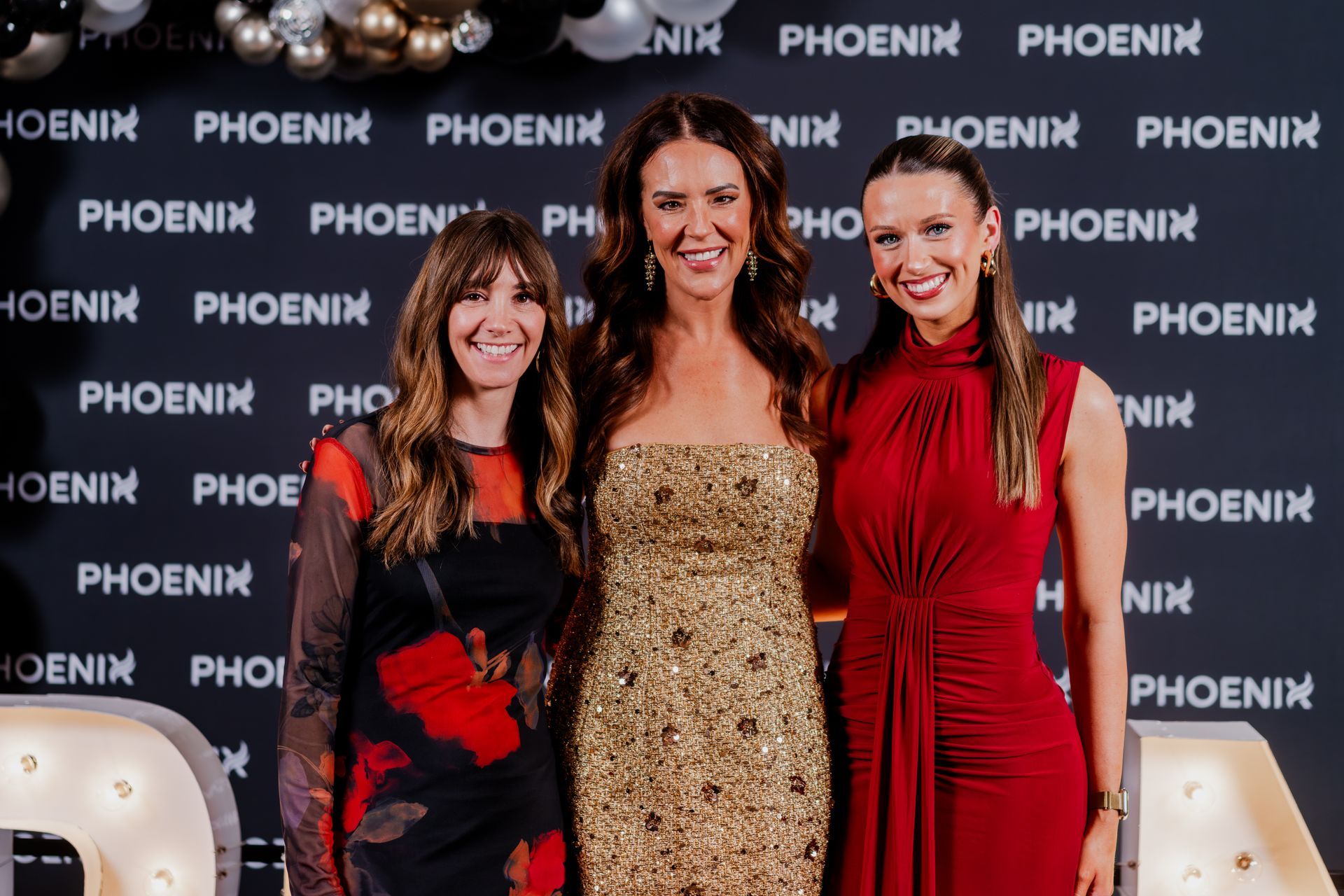 Three people smiling in elegant evening attire, standing together in front of a branded backdrop for a Phoenix event.