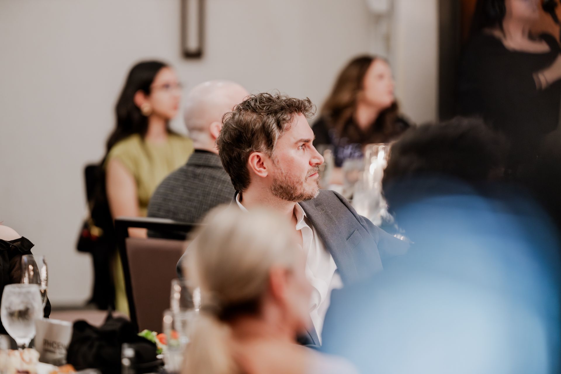 A group of people seated at a dinner event, focused on a speaker, with a man in a blazer in the foreground.