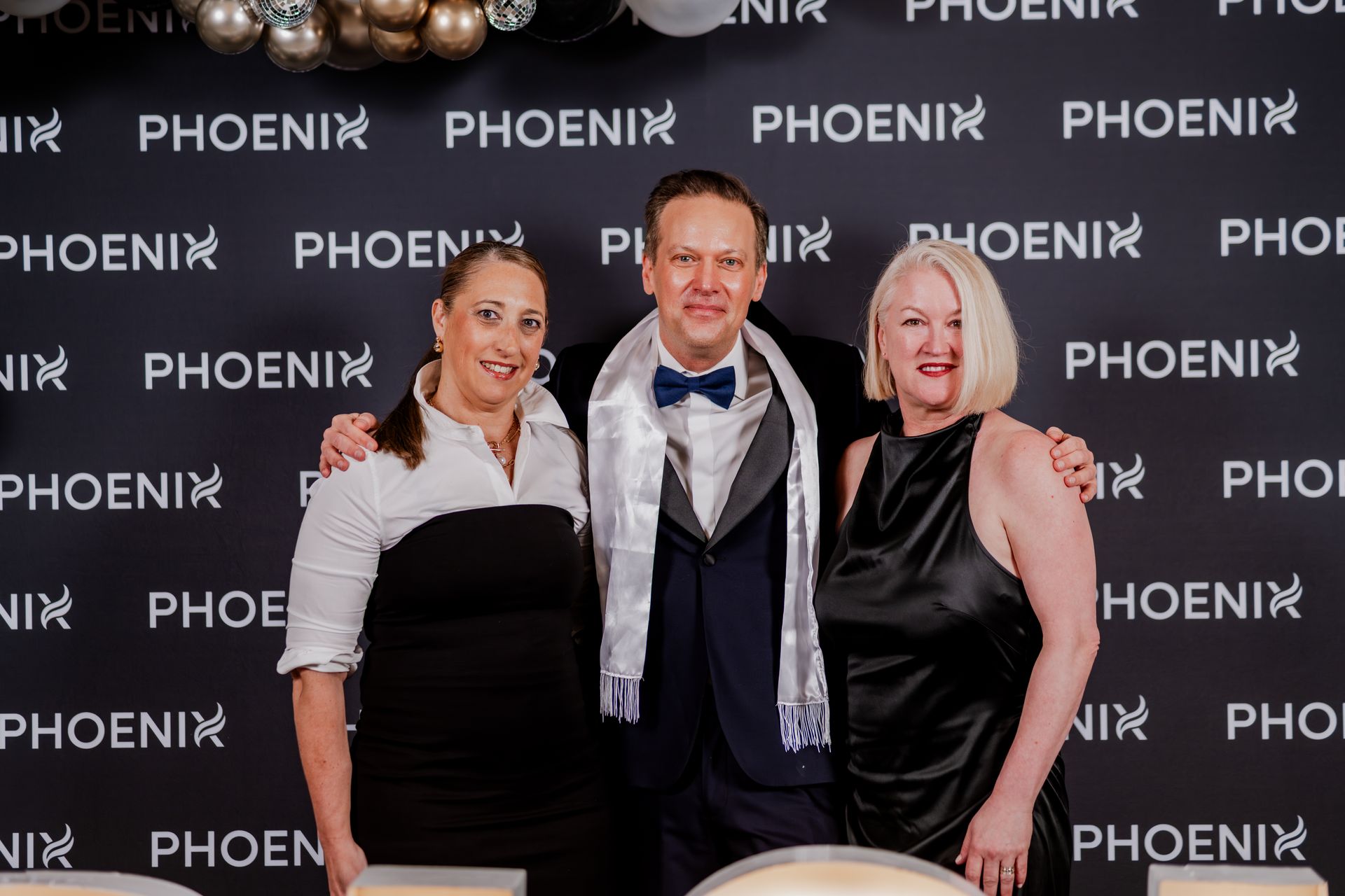 Three people standing together and smiling in front of a black step-and-repeat backdrop featuring the word PHOENIX.