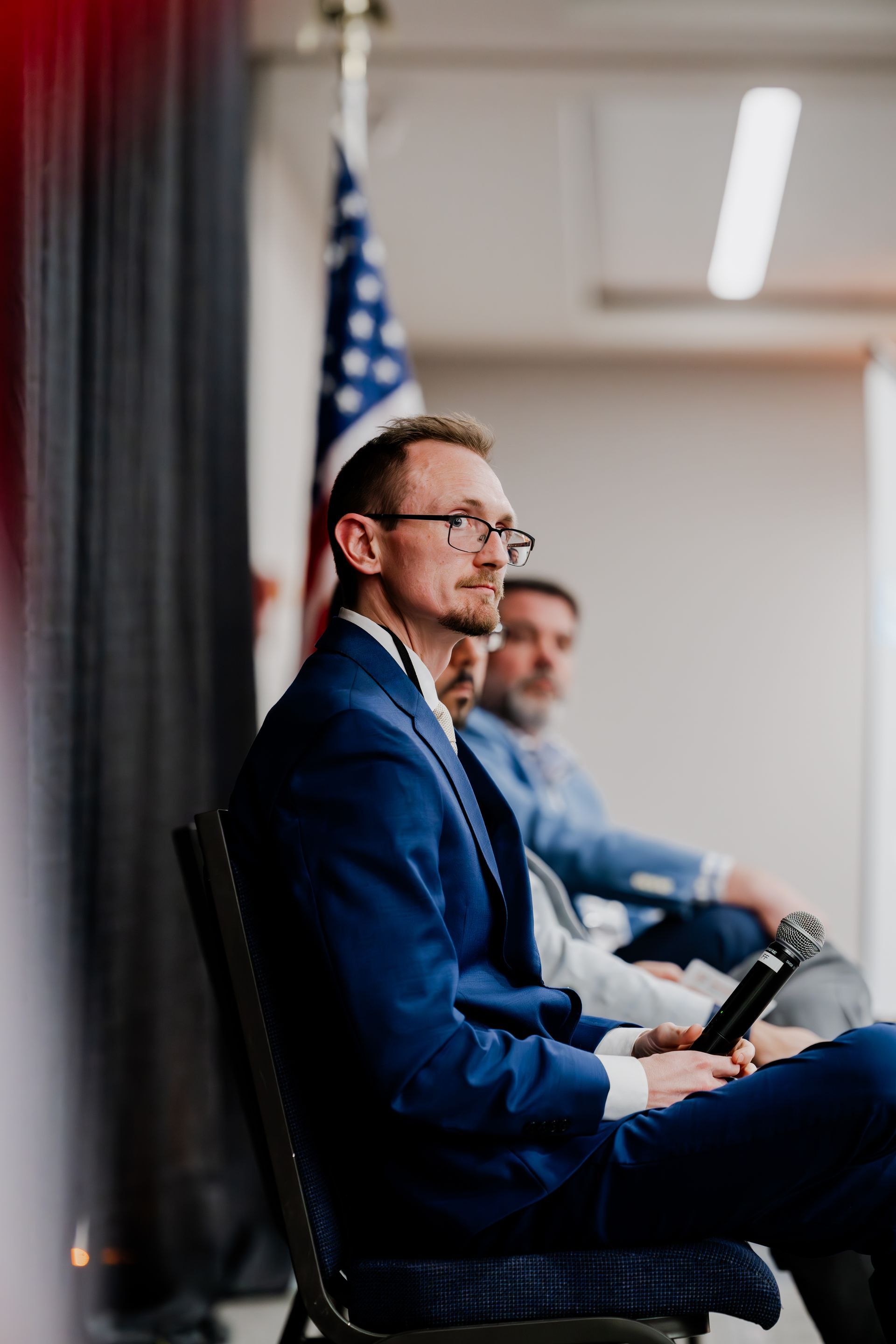 Two men in business suits sit on chairs during an indoor event with an American flag displayed in the background.