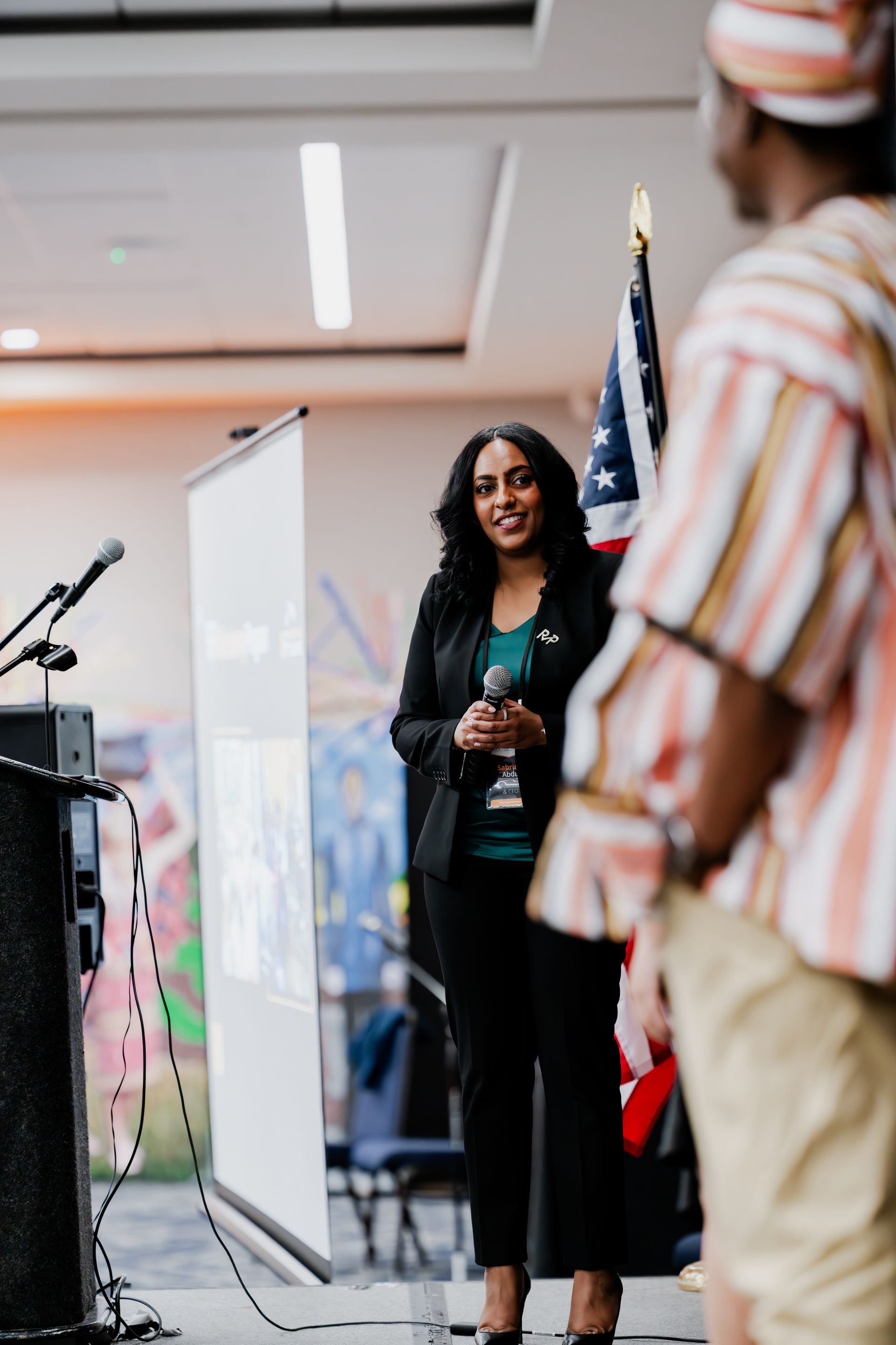 Woman in a black suit speaks at a podium; another person stands nearby. American flag in background.