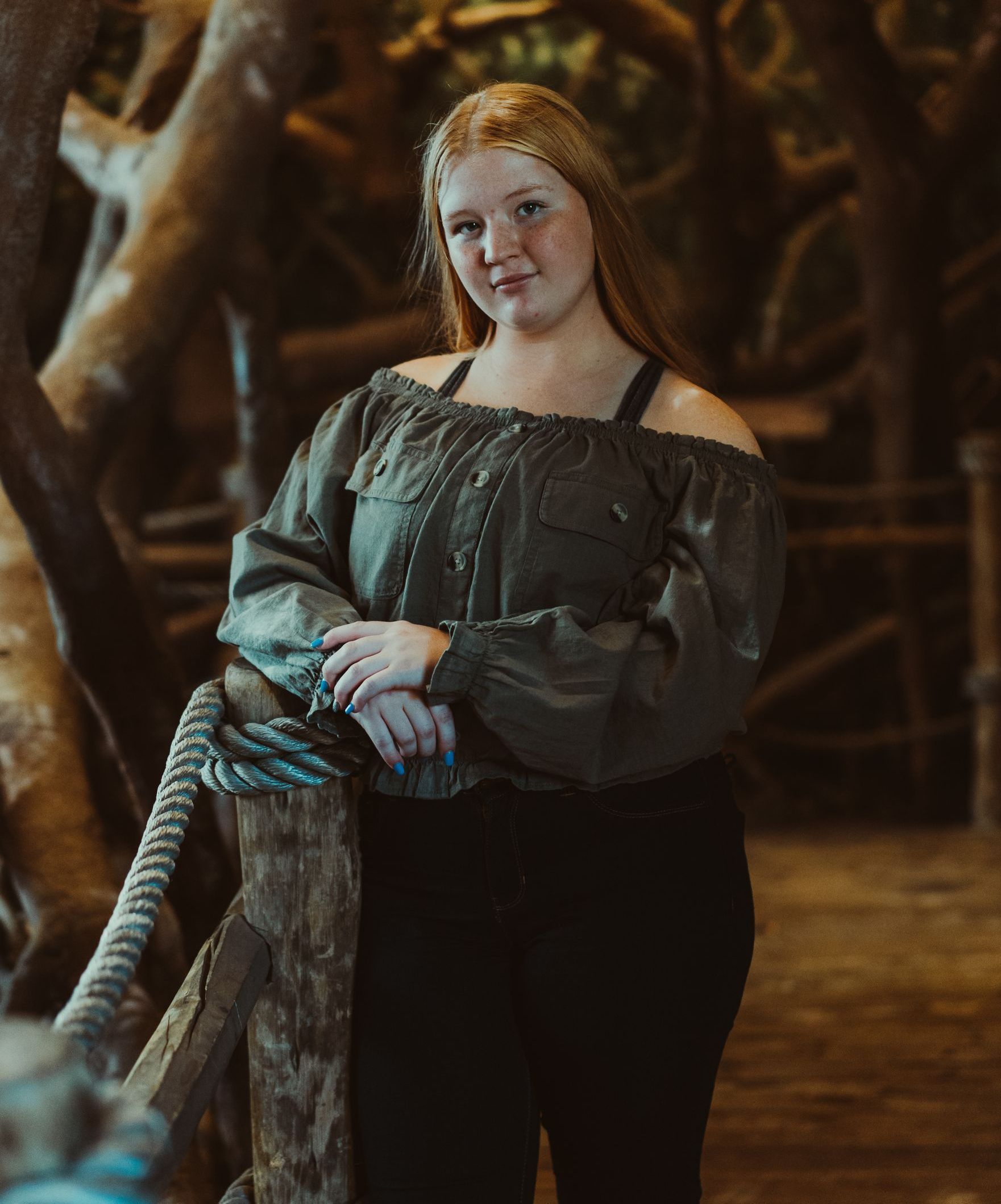 high school senior in a green off the shoulder top is standing next to a tree .