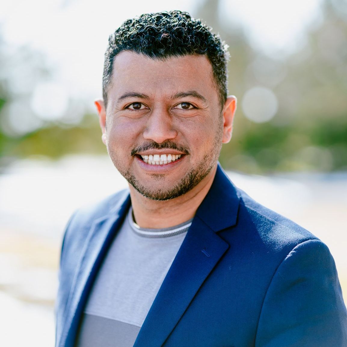 Headshot of a man in a blue suit and gray shirt is smiling for the camera .