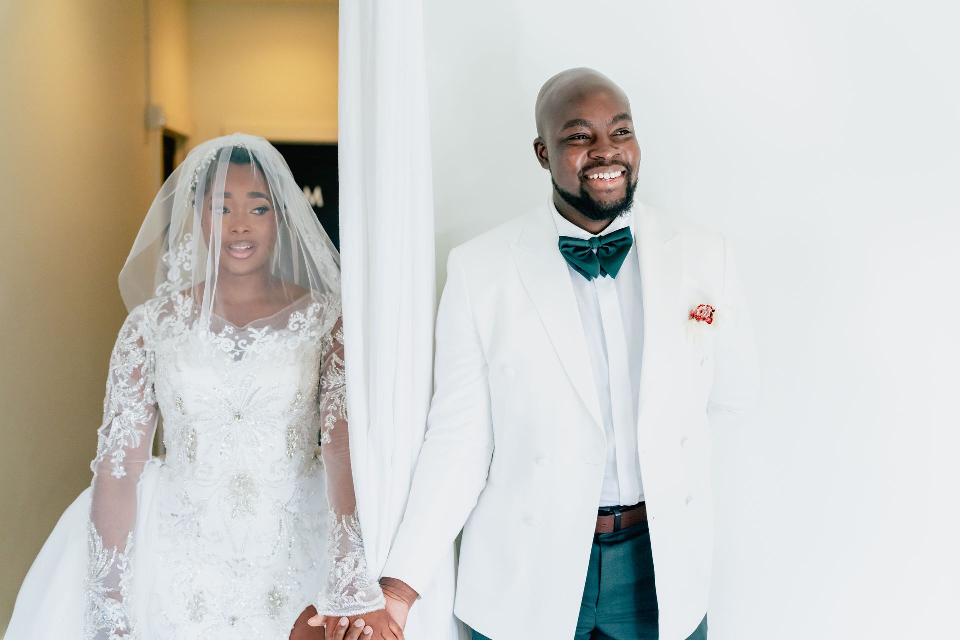 Bride and groom hold hands, smiling. Bride wears white gown and veil, groom white suit with bow tie.